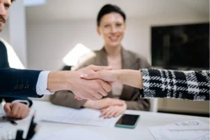 Two professionals shaking hands across a desk with a smiling colleague witnessing the agreement